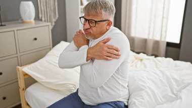 Orta yaşlı bir adam iyi aydınlatılmış bir yatak odasında yatağında otururken omuz ağrısı çekiyor.