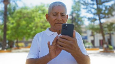 Beyaz saçlı İspanyol adam güneşli bir günde şehir parkında akıllı telefon kullanıyor..
