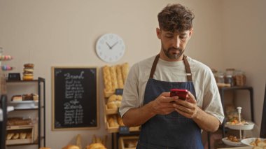 Genç adam fırında akıllı telefon kullanıyor, ekmek ve pastalarla çevrili, önlük takıyor, içeride duruyor, raflarla kaplı ve arka planda bir duvar saati var..