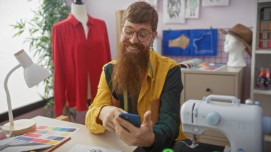 Sakallı kızıl saçlı bir adam, elinde akıllı bir telefonla, renkli bir terzi dükkanında, dikiş ekipmanları ve kumaş örnekleriyle gülümsüyor..