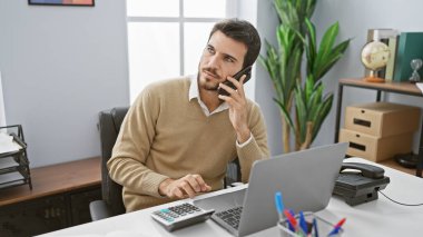 Ofiste çalışan sakallı genç bir İspanyol. Laptop, hesap makinesi ve akıllı telefon kullanıyor..