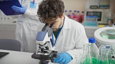 Laboratuvarda mikroskop kullanan bir adam bilimsel ekipman ve elinde bir pano tutan bir meslektaşı var..