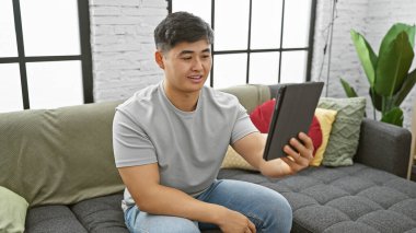 Genç Asyalı bir adam, modern bir apartman dairesinde, kanepenin üzerinde rastgele tablet kullanıyor.