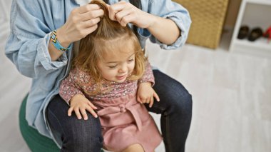 Mavi gömlekli gülümseyen bir anne pembe elbiseli, güzel ışıklandırılmış bir soyunma odasında oturan mutlu kızının saçlarını örüyor..