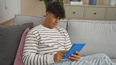 Yakışıklı genç İspanyol bir adam oturma odasında oturuyor, tablet kullanıyor ve çizgili bir kazak giyiyor, odaklanmış ve rahat görünüyor, rahat, modern bir iç mekan ile çevrili..