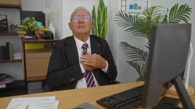 Takım elbiseli kıdemli, gri saçlı, beyaz bir adam ofisindeki masasında oturmuş bilgisayarın yanında göğsünü sıkıyor ve ofis bitkileri ve malzemelerle çevrili..
