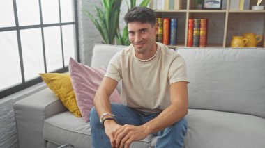 Güzel dekore edilmiş bir oturma odasındaki kanepede otururken gelişigüzel gülümseyen yakışıklı İspanyol adam.