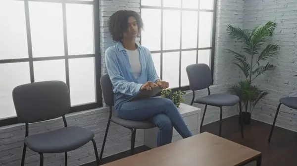 stock image A beautiful young african american woman with curly hair sits in a modern indoor waiting room or lobby featuring stylish chairs and a windowed backdrop.