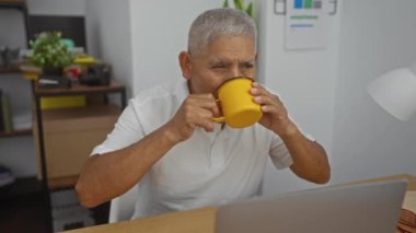 Ofisinde bitki ve ofis malzemeleriyle çevrili bir laptopta çalışırken sarı bardakta içkinin tadını çıkaran olgun İspanyol bir adam..