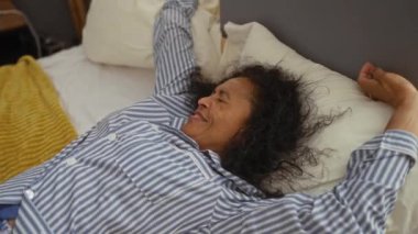 Yatakta çizgili pijama giyen bir kadın, sabah rutinini içeren bir ev yatak odası ortamında.