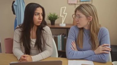 Evde dizüstü bilgisayar ve belgelerle oturan iki kadın, modern bir oturma odasında profesyonel ya da kişisel bir tartışmayı canlandırıyorlar..