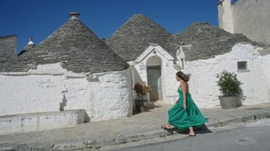 İtalya 'nın Alberobello kentindeki büyüleyici trulli evlerde açık mavi gökyüzünün altında yeşil elbiseli genç bir İspanyol kadın yürüyor..