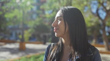 Gülümseyen genç bir kadın, arka planda bulanık yeşil ağaçlarla güneşli bir parkın tadını çıkarıyor..