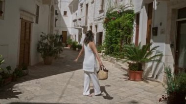 Beyaz elbiseli genç bir İspanyol kadın neşeli bir şekilde Locorotondo, Puglia, İtalya sokaklarında yürüyor. Beyaz binalar ve canlı bitkilerle çevrili..