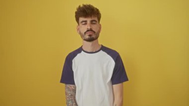 Genç İspanyol bir adam güvenle ayakta duruyor, çenesi elinde dinleniyor, kolları çapraz, izole edilmiş sarı bir arka planda pozitif bir gülümsemeyle kameraya bakıyor.