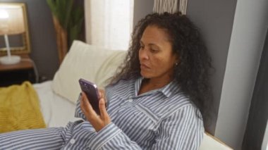 Yatak odasında uzanmış, çizgili pijama ve lamba komodinin üzerinde ev dekorasyonuyla telefonda konuşan bir kadın..