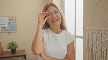 Gözlüklü, gülümseyen olgun bir kadın dekoratif makromla rahat bir oturma odasında rahatça poz veriyor..