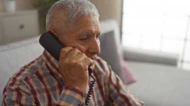 Beyaz saçlı, yaşlı İspanyol bir adam oturma odasında otururken telefonla konuşuyor..