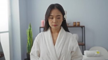 Beyaz cüppeli, saçları çiçekli Asyalı bir kadın. Huzurlu bir spa odasında duruyor. Raflarda çiçekler ve mumlar var..
