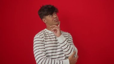 Çizgili tişört giyen genç İspanyol adam mutlu bir fikirle parmağını kaldırıyor ve izole edilmiş kırmızı arkaplanda başarı için bir numaralı jest.