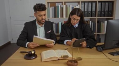 Kadın ve erkek büroda bir tablet, hukuk kitapları, tokmak ve masanın üzerindeki teraziyle belgeleri tartışıyor.