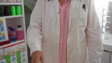 Beyaz önlüklü, çapraz kollu bir kadın eczacı ilaç ve ürünlerle dolu bir eczanede duruyor.