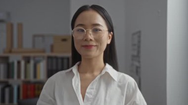 Çin 'de bir ofiste gözlük takan genç Asyalı bir kadın, arka planda kitap ve klasörleri olan kapalı bir iş yerinde güvenle gülümsüyor.