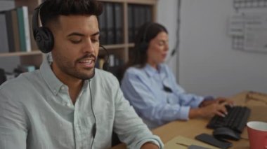 Kadın ve erkek, ofiste birlikte çalışıyor. Kulaklık takıyor, kapalı bir işyerindeki görevlerine odaklanıyorlar.