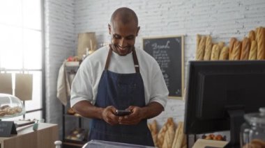Bir fırında gülümseyen genç adam tezgahın arkasında telefonunu kontrol ederken arka planda ekmek ve hamur işleri sergileniyor.