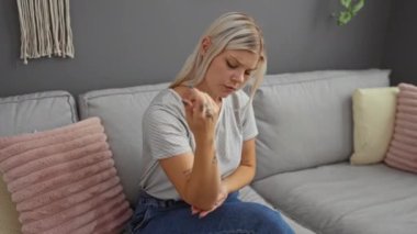 Kolunu muayene eden bir kadın modern bir oturma odasında gri duvarları ve pembe minderleri olan