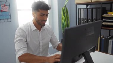 Ofis ortamında çalışan, beyaz gömlekli, bilgisayarlı bir masada oturan, etrafı dosya, bitki ve motivasyon notlarıyla çevrili genç İspanyol bir adam.