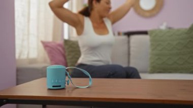 Oturma odasındaki kanepede oturmuş, mavi bluetooth hoparlörüyle ahşap masada dans eden bir kadın.
