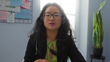 Kulaklık takan kadın ofiste oturuyor, kapalı gözlerle, büyük gözlüklerle ve koyu uzun saçlarla iletişim kuruyor, arka plandaki duvarda renkli notlarla, odak ve konsantrasyonu vurguluyor.