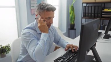Ofiste çalışan orta yaşlı bir adam, akıllı bir telefonla konuşuyor ve masasındaki bilgisayar klavyesinde daktilo kullanıyor. İç dekorasyon ve ofis ekipmanlarıyla profesyonel bir iş yeri ayarını gösteriyor.