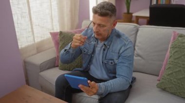 Orta yaşlı bir adam rahat oturma odasında tablet kullanıyor, gözlük takıyor, kot ceket giyiyor, koltukta oturuyor, modern iç mekan, bej perdeler, yeşil yastıklar, pembe duvarlar, odaklı ifade