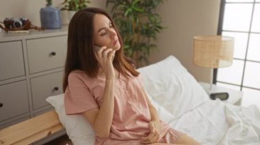 Yatak odasında, çiçekli ve şifoniyerli bir odada oturan bir kadın telefonda konuşuyor.