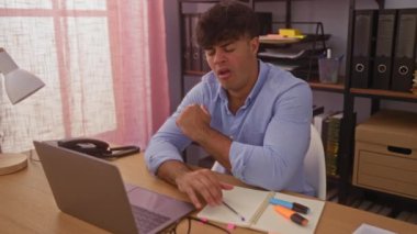 Genç, İspanyol, modern ofis çalışma alanında esneyen, elinde laptop ve ofis malzemeleriyle bir masada oturan, çalışma saatlerinde yorgun görünen bir adam..