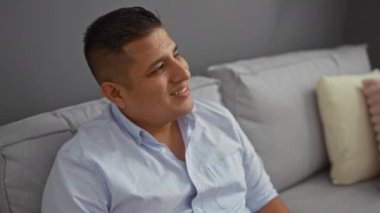 Oturma odasındaki kanepede oturan genç İspanyol adam gülümsüyor ve yan tarafa bakıyor.