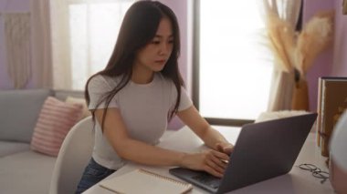 Genç bir kadın rahat bir ev ortamında dizüstü bilgisayarla çalışırken bir not defterine not alıyor.