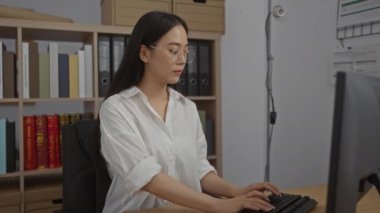 Ofisteki bir masada çalışan genç bir kadın, kitaplarla ve dosyalarla dolu raflarla çevrili, gözlüklü ve beyaz gömlekli.