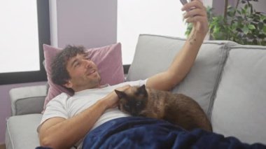 Genç bir İspanyol adam, modern bir oturma odasında kedisiyle rahat bir kanepede rahatlarken selfie çekiyor..