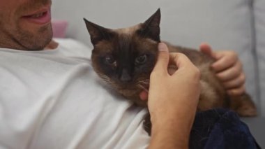 Kapalı bir ortamda, genç bir adam bir siyam kedisini nazikçe okşar, şefkat ve dostluk anı gösterir..