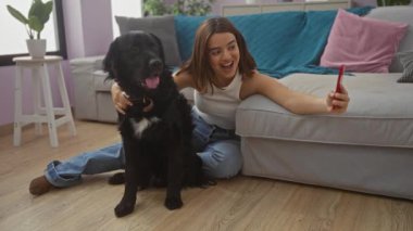 Genç bir kadın, siyah labrador köpeğiyle rahat bir oturma odasında selfie çekiyor. Kapalı bir ortamda neşeli bağlarını sergiliyor..