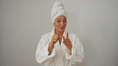 Havlu ve bornoz giymiş orta yaşlı bir kadın, mutlu, komik bir ifadeyle parmaklarını kameraya doğrultuyor, izole beyaz arka planda güzel bir enerji yayıyor.