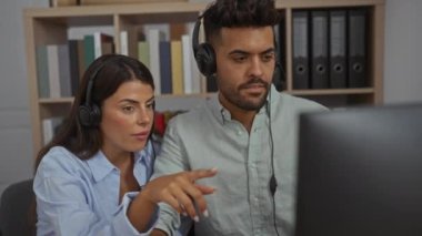Kadın ve erkek, ofiste birlikte çalışıp kulaklık takıyor ve bilgisayar ekranına odaklanıyor..