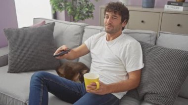 Genç bir İspanyol adam oturma odasında oturuyor, sarı bir kupa tutarken bir kediyi okşuyor, ve beyaz gömlek ve kot pantolon giyiyor..