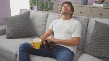 Rahat bir adam, bir kanepede siam kedisi ve rahat bir oturma odasında bir kupayla bir dakikalık sükunetin tadını çıkarır..