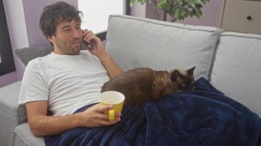 Rahatlamış bir adam telefonda konuşur, evde bir kanepede siyam kedisiyle oturur, elinde bir kupayla..