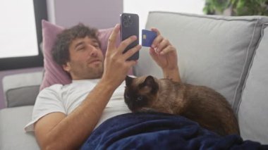 Elinde kredi kartı tutan ve yanında kanepede oturan siyam kedisi olan akıllı telefon kullanan gülümseyen bir adam.