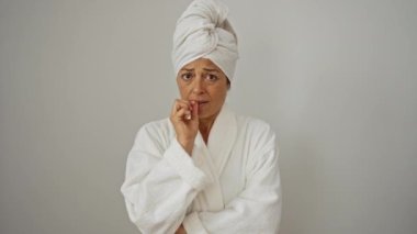 Gergin orta yaşlı kadın, bornoz ve havlu giymiş, anksiyete ve stresten tırnaklarını yiyor, beyaz arka planda soyutlanmış.
